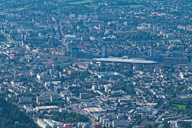 Eine Luftaufnahme einer belebten Stadt mit hohen, bunten Gebäuden, saftig grünen Bäumen, gewundenen Straßen und leuchtend grünem Gras, mit Menschen, die spazieren gehen und Autos fahren.