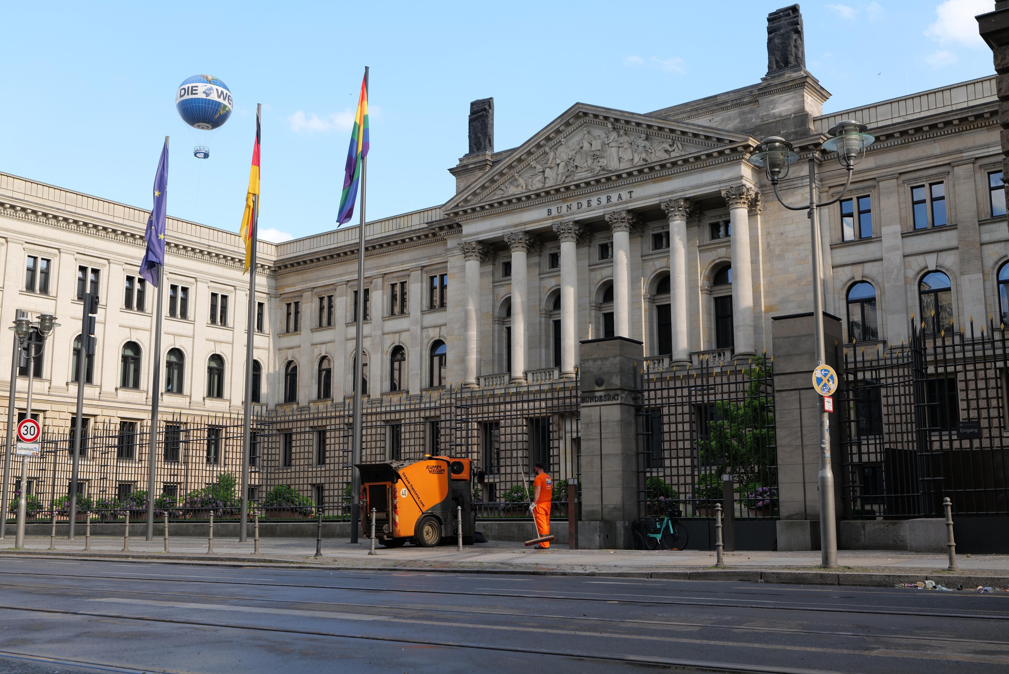 Large modern building with many windows, identified as the Bundestag in Berlin, Germany, surrounded by street infrastructure, pedestrians, and trees under a cloudy sky.