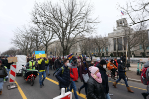 A large group of people participating in a protest march in Washington, D.C. on January 21, 2020, walking down a street with placards, banners, and bicycles, against a backdrop of sign boards, trees, and a clear blue sky.