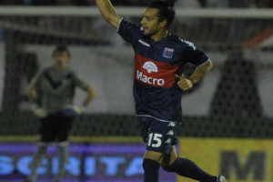 Ein Fussballspieler liegt auf dem Boden und feiert ein Tor, mit einer stehenden Person und einer Texttafel im Hintergrund, während eine jubelnde Menge in der Ferne unscharf ist.