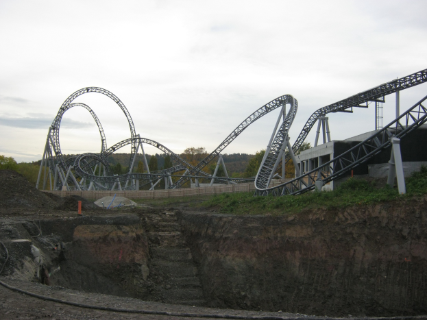 Eine Achterbahn fährt einen Hügel mit einer Brücke, Bäumen und einem klaren blauen Himmel im Hintergrund und einer Eisenbahnschiene, die sich am Fuße des Hügels windet.
