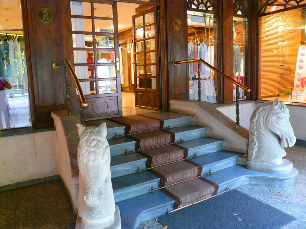 Eingang zu einem Geschäft mit zwei Pferdestatuen auf beiden Seiten der Treppe, Teppichboden, Glastüren, Schaufensterpuppen in Kleidern und Deckenbeleuchtung.