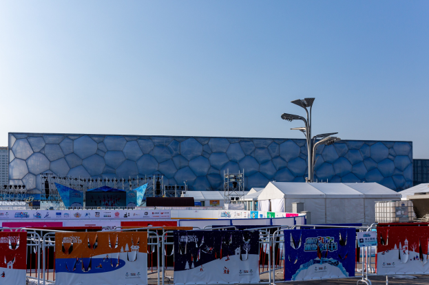 Olympiastadion in Sotschi, Russland, mit Bannern, Geländern, Zelten, Laternenmasten und Gebäuden im Vordergrund unter einem klaren blauen Himmel.
