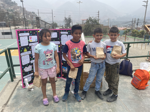 Gruppe von Kindern, die Papierbeutel auf einem Basketballplatz halten, mit Beuteln und Gegenständen auf dem Boden, einer Tafel mit Text links und einem Hintergrund mit Zaun, Pfosten, Straßenlaternen, Bäumen, Gebäuden, Hügeln und Himmel.