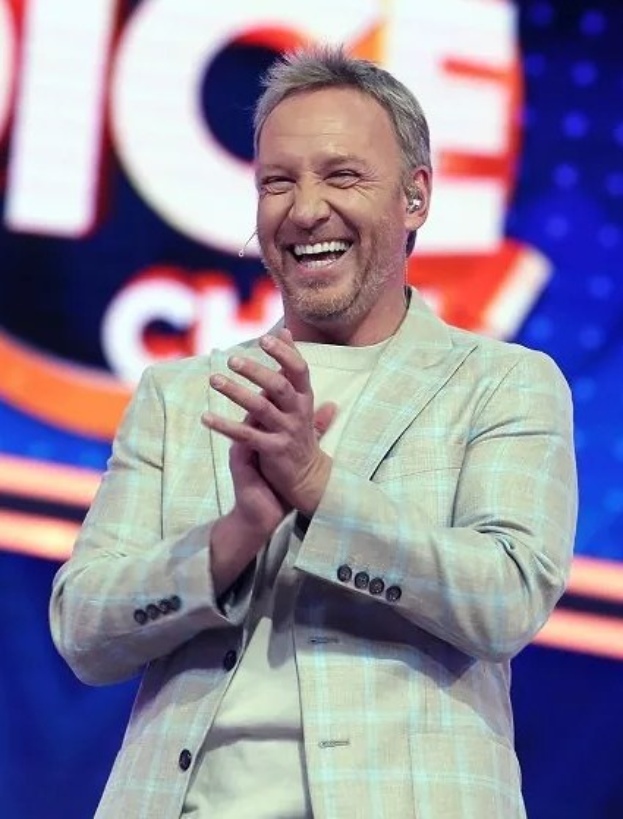 A man in a suit clapping on a stage with "The Voice" logo visible in the background.
