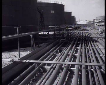 Black and white photo of a large group of pipes arranged in a neat line at a factory, with buildings in the background and a clear sky above, with text at the top and bottom of the image.