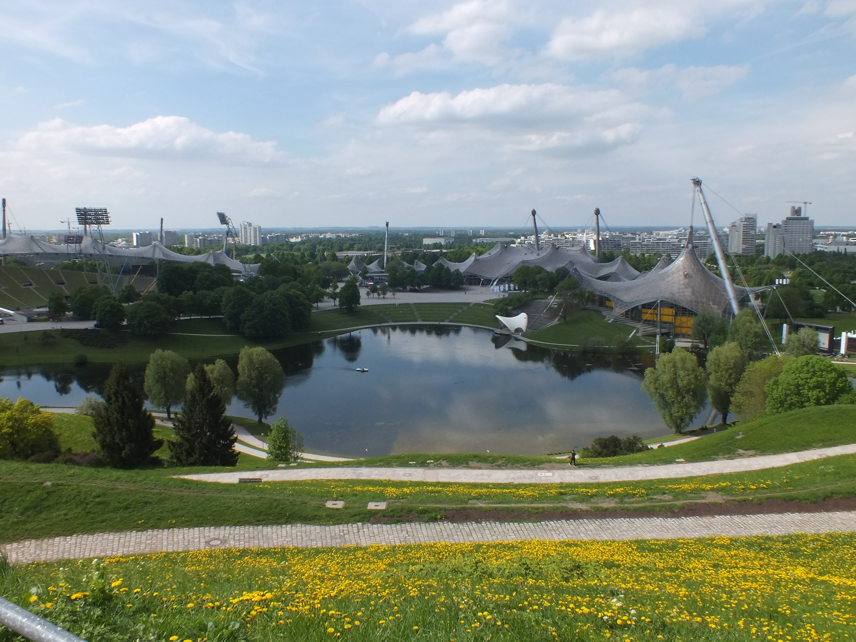 View of Olympic Stadium from a hilltop, with a pond surrounded by greenery and yellow flowers in the foreground and buildings against a clear blue sky in the background.