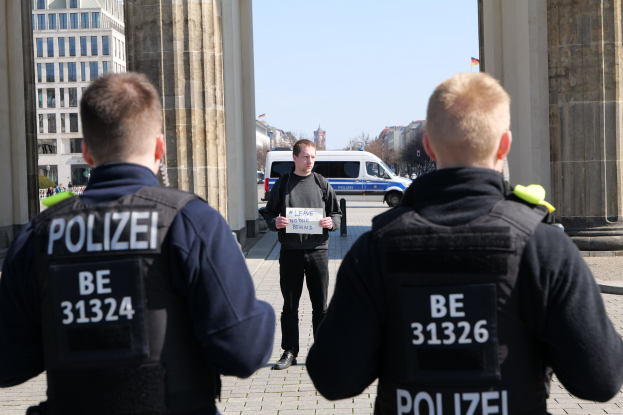 Three police officers stand in front of a building, with a man holding a paper between them, flanked by pillars, as vehicles, people, trees, and buildings fill the background under the sky.