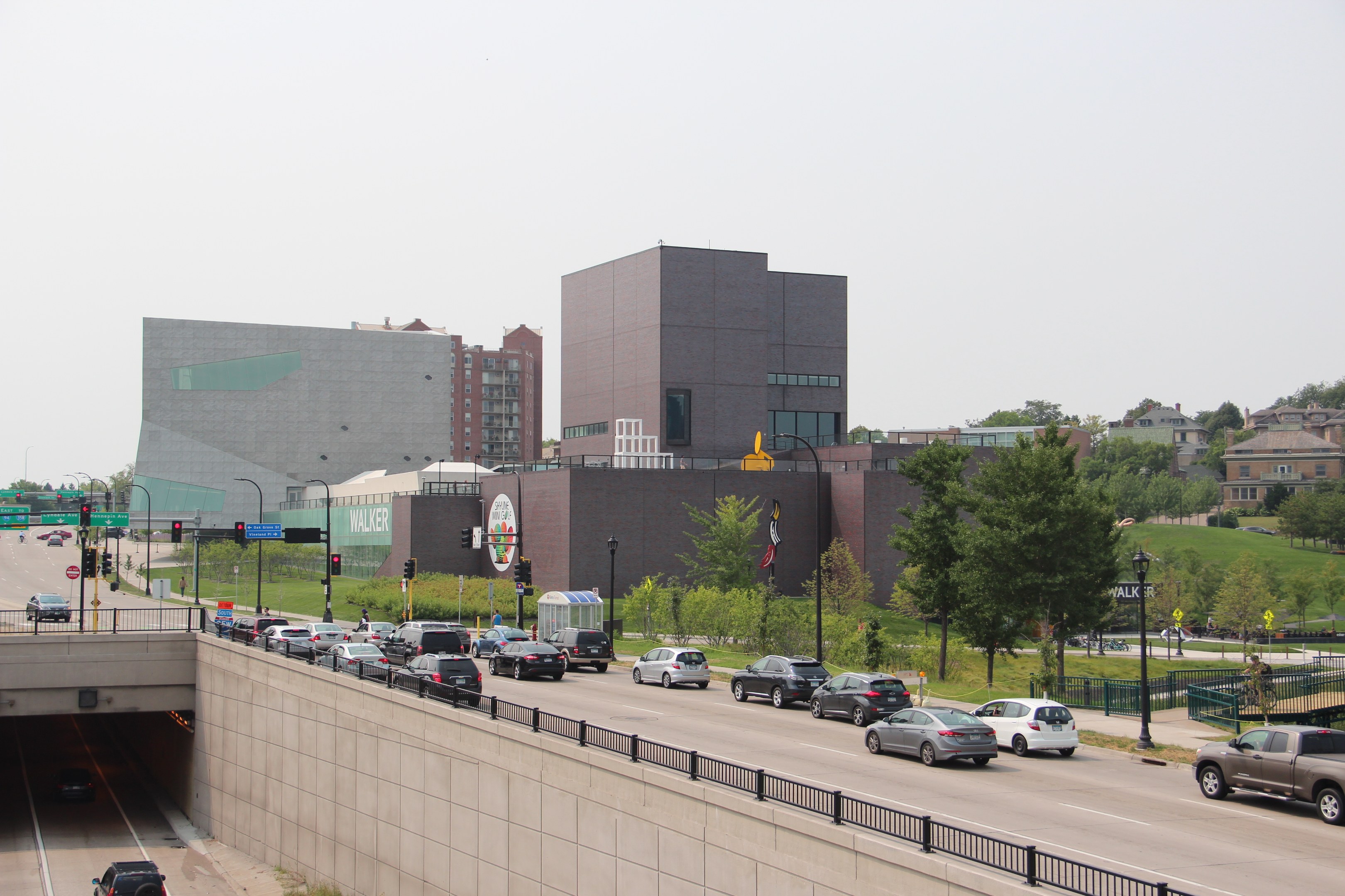 City street with moving cars, a bridge with railings, light poles, traffic signals, trees, buildings with windows, and a sky background.