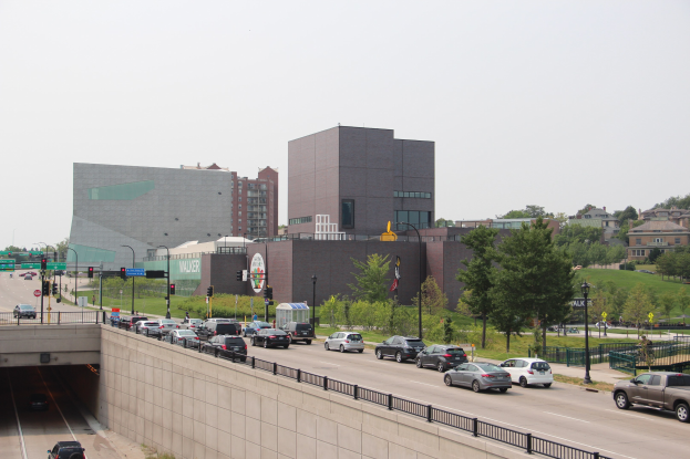 City street with moving cars, a bridge with railings, light poles, traffic signals, trees, buildings with windows, and a sky background.