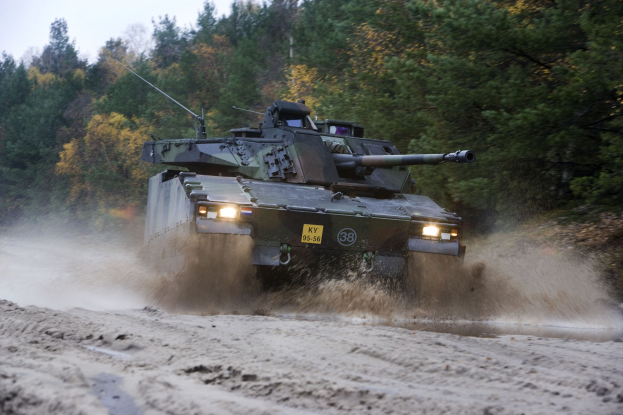 Ein militärischer Panzer mit einer montierten Kanone fährt eine Schotterstraße entlang, die von Bäumen gesäumt ist und einen klaren Himmel hat.