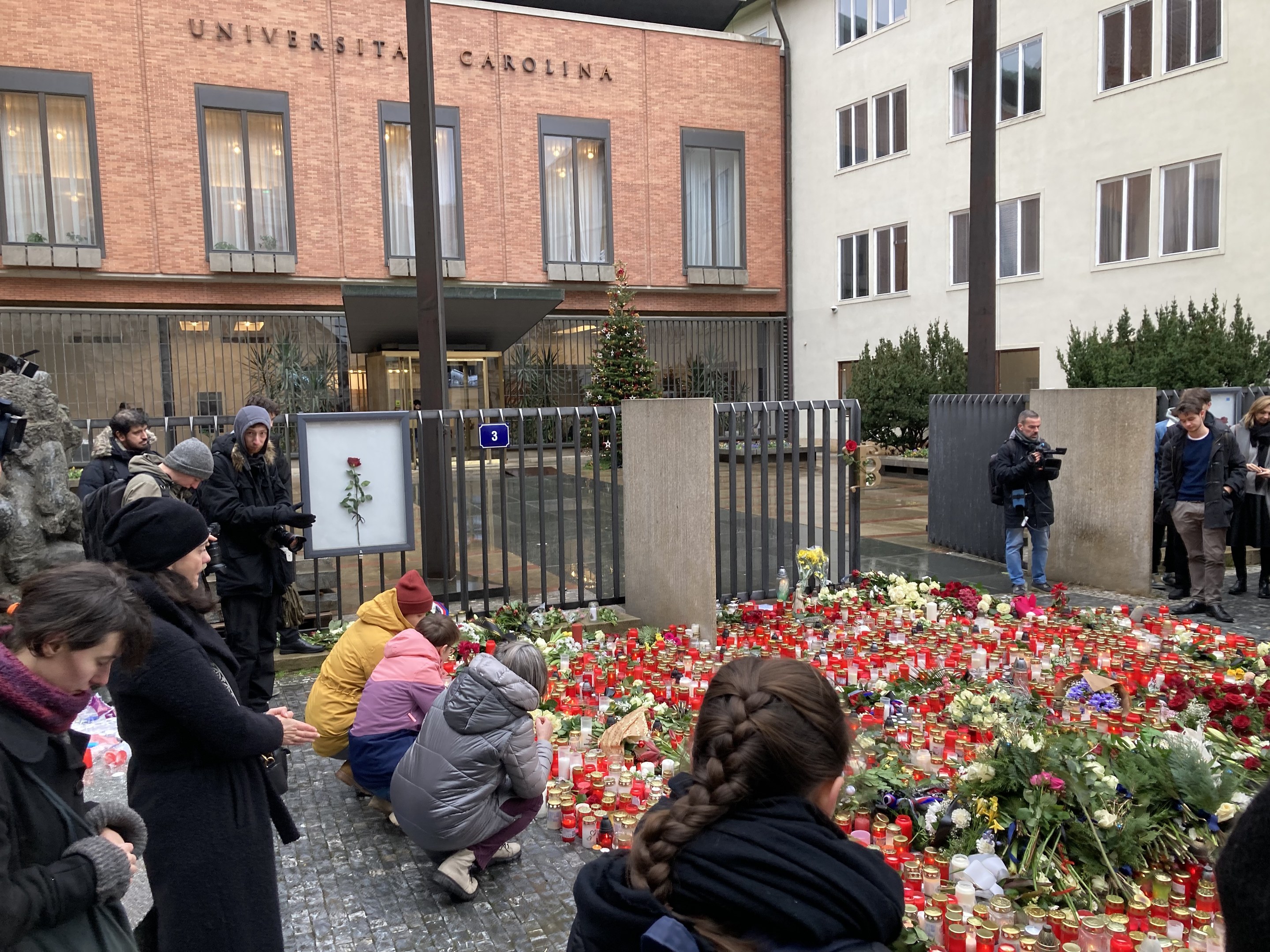 A group of people gather around flowers and candles placed on the ground outside a building, some wearing caps and holding cameras or bouquets, with buildings, trees, and railings in the background.