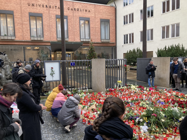 A group of people gather around flowers and candles placed on the ground outside a building, some wearing caps and holding cameras or bouquets, with buildings, trees, and railings in the background.