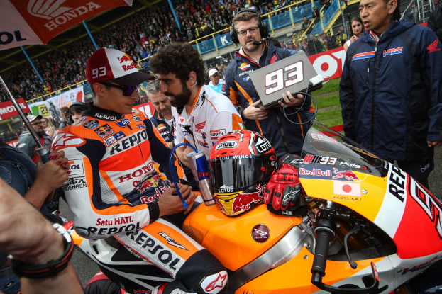 Eine Gruppe von Menschen um ein Motorrad auf einer Rennstrecke versammelt, mit einer Person in einem weißen, orangen und roten Kleid und Helm vorn und einer anderen Person mit einem Schirm links; Zuschauer füllen das Stadion und die grasbewachsenen Bereiche im Hintergrund.