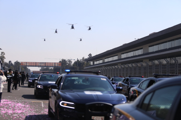 Polizeifahrzeuge vor einem Gebäude mit Menschen auf der linken Seite, Bäumen und einer Brücke im Hintergrund sowie vier Hubschraubern am Himmel.