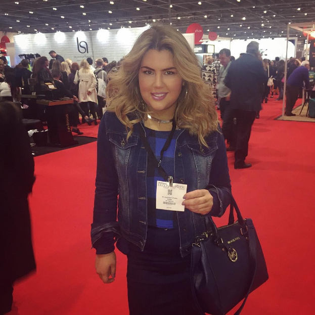 Eine Frau steht auf einem roten Teppich, lächelt und hält eine Handtasche, mit Menschen, Lichtern und Schildern im Hintergrund bei einer Modenschau.