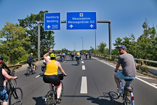 Group of cyclists wearing helmets riding down a road with trees on one side and a railing on the other, light poles in the background, and a clear blue sky, with a sign at the top indicating a cycling tour in Hamburg.
