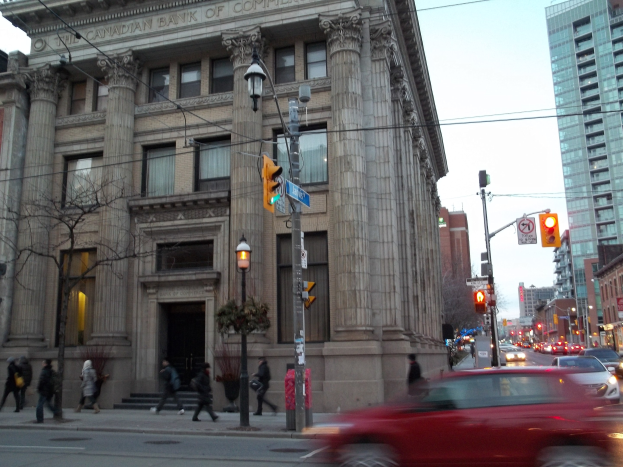 Eine belebte Stadtstraße mit Fahrzeugen, Fußgängern, Verkehrsampeln, Schildern, Bäumen und Gebäuden unter einem sichtbaren Himmel, mit einem roten Auto im Vordergrund und einem großen Gebäude.