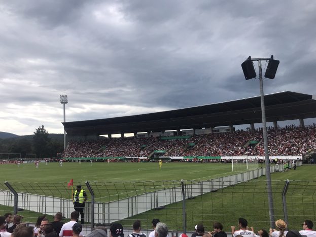 Große Zuschauermenge bei einem Fußballspiel in einem Stadion mit beleuchtetem Feld, umgeben von einem Zaun, Bäumen, Hügeln und bewölktem Himmel.