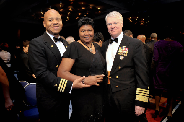 Zwei Männer in schwarzen Blazern und weißen Hemden stehen neben einer Frau in einem schwarzen Kleid, die eine Auszeichnung hält, auf einem roten Teppich, während andere im Hintergrund sitzen und stehen, unter beleuchteten Decken.