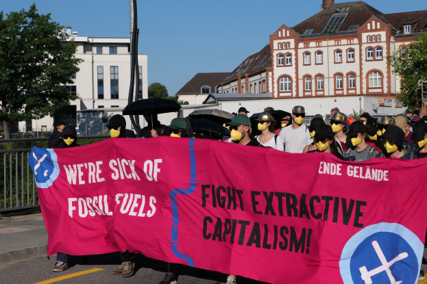 Eine Gruppe von Menschen mit Masken, die eine rosa Schrifttafel mit der Aufschrift "Wir haben genug von fossilen Brennstoffen, bekämpft ausbeuterisches Kapitalismus" tragen, auf einer Straße mit Schirmen, Gebäuden, Bäumen, einem Pfahl und einem klaren blauen Himmel im Hintergrund.