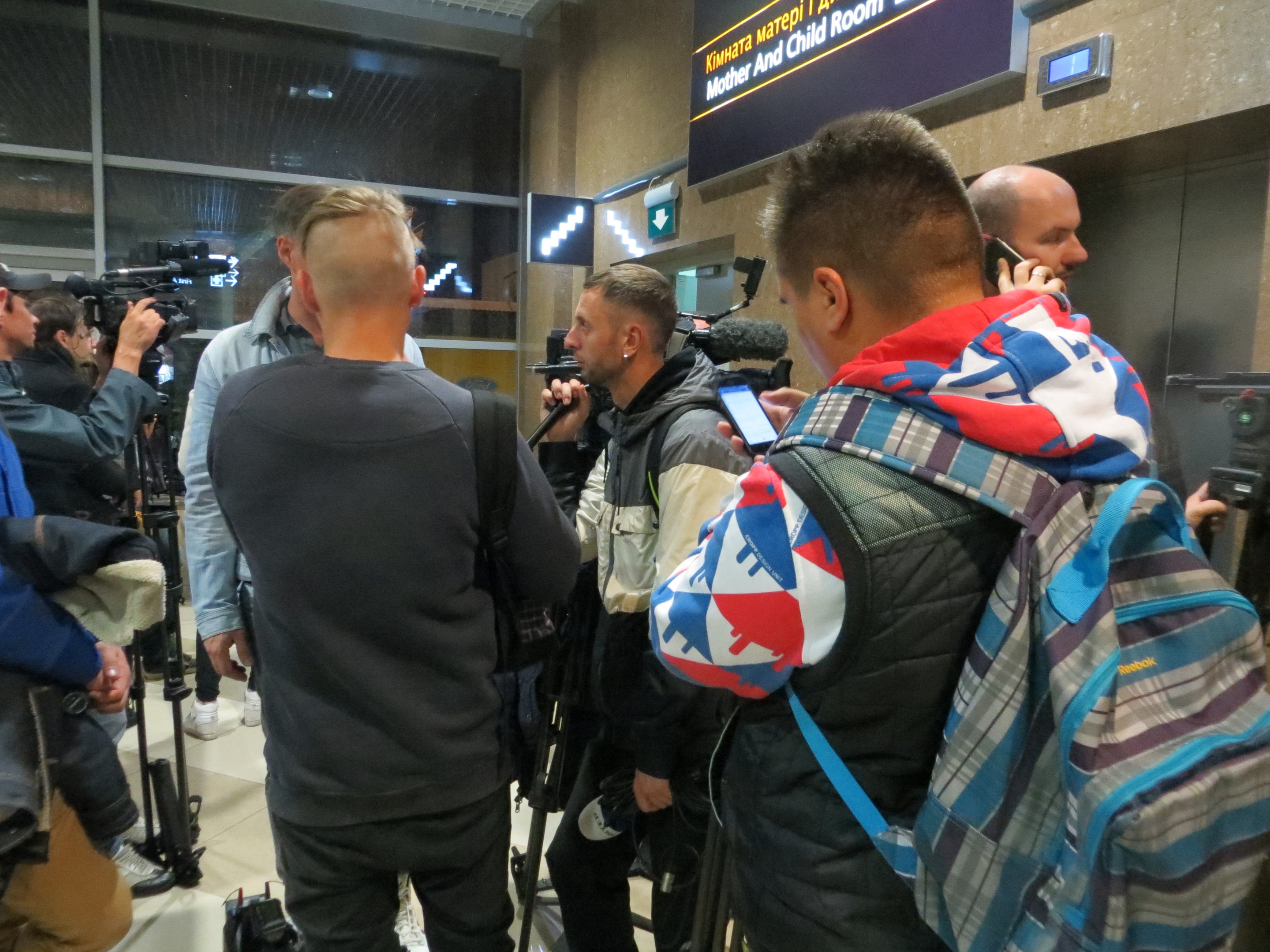 Group of journalists at an airport press conference, some holding cameras and phones, with a text board and overhead lights in the background.