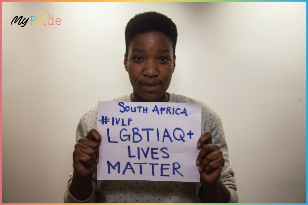 A woman in a white shirt holds up a sign reading "LGBTQ+ Lives Matter" in front of a wall, her expression conveying solidarity.
