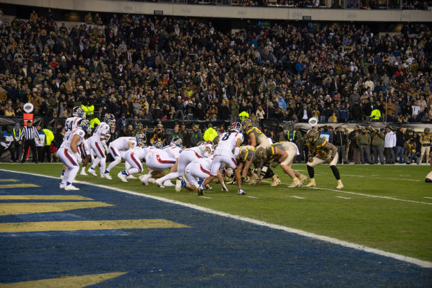 Ein Fütbolspiel im Gange mit Spielern in Sportkleidung und Helmen auf dem Feld, Zuschauern, Geländern und Tafeln mit Text im Hintergrund.