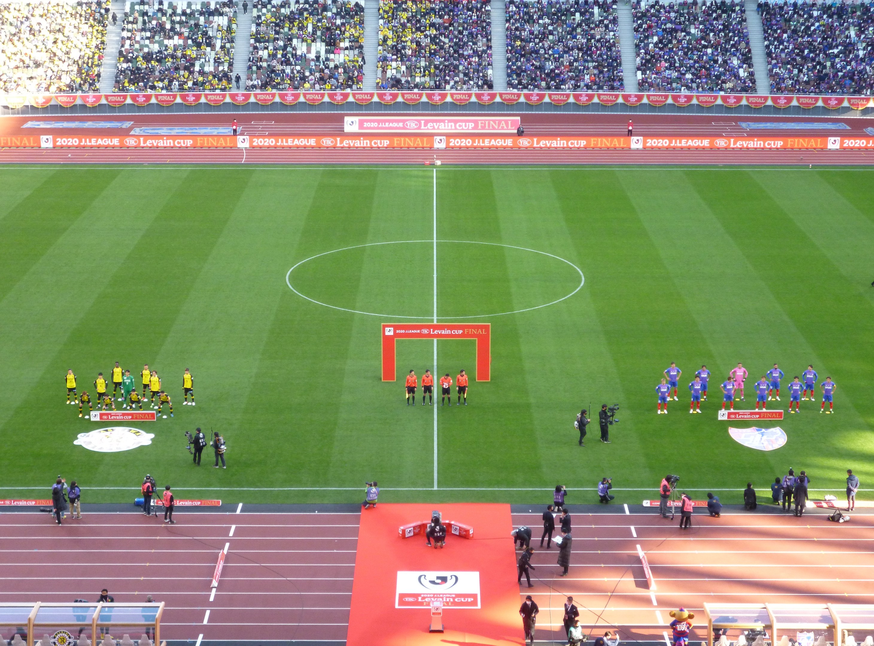 Eine Gruppe von Menschen steht auf einem grünen Fußballfeld in einem Stadion, das von sitzenden Zuschauern erfüllt ist, mit Werbetafeln, die um das Feld herum sichtbar sind.