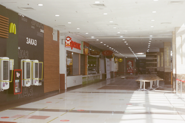 Interior of a fast food restaurant with tables, chairs, a wall-mounted menu and machines, overhead lighting, and a glass exterior wall.