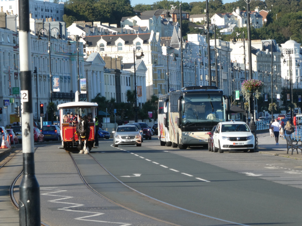 Eine belebte Stadtstraße mit hohen Gebäuden, Fahrzeugen, Fußgängern, Verkehrsampeln, Laternenmasten, Bäumen und einem klaren blauen Himmel.