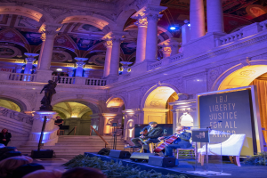Group of people seated on a stage with musicians, a podium, and a board during the Liberty Justice for All Award Ceremony in New York City.
