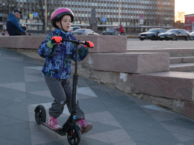 Ein junger Junge fährt mit einem Roller auf einem Gehweg, trägt einen Helm und Handschuhe, mit verschiedenen städtischen Elementen und einem klaren blauen Himmel im Hintergrund.