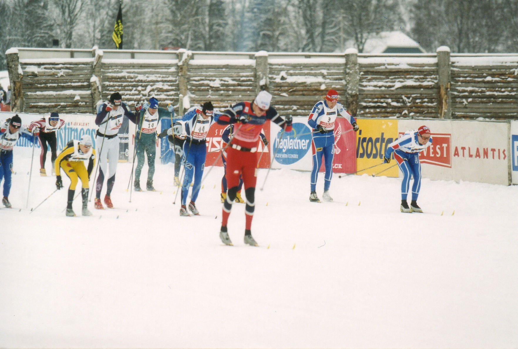 Gruppe von Menschen, die eine schneebedeckte Piste mit Ski-Stöcken hinunterfahren, mit Bannern, einem hölzernen Zaun, Fahnen, Bäumen, Häusern und einem klaren blauen Himmel im Hintergrund.