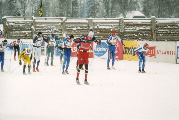 Gruppe von Menschen, die eine schneebedeckte Piste mit Ski-Stöcken hinunterfahren, mit Bannern, einem hölzernen Zaun, Fahnen, Bäumen, Häusern und einem klaren blauen Himmel im Hintergrund.
