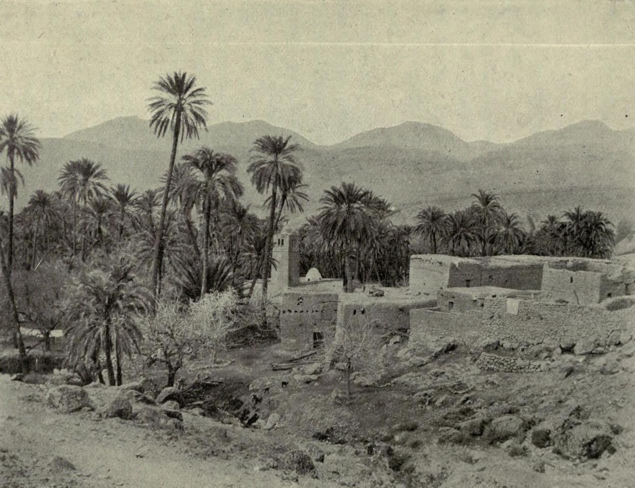 Old black and white photo of a desert village with houses, palm trees, vegetation, rocks, and mountains in the background, with text at the bottom.