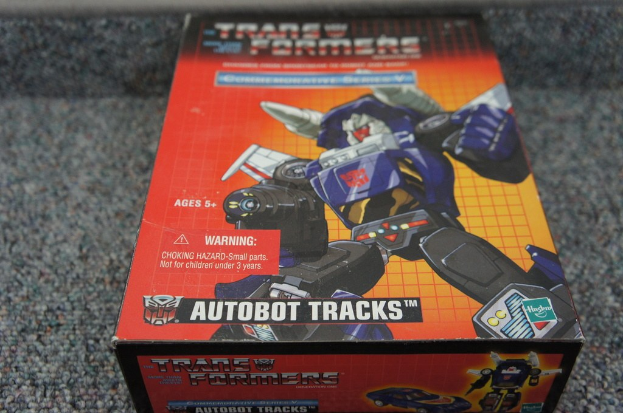 Ein roter, oranger und blauer Autobot Tracks Roboterkasten mit "AUTOBOT TRACKS"-Text oben drauf, vor einem unscharfen schwarzen Hintergrund.