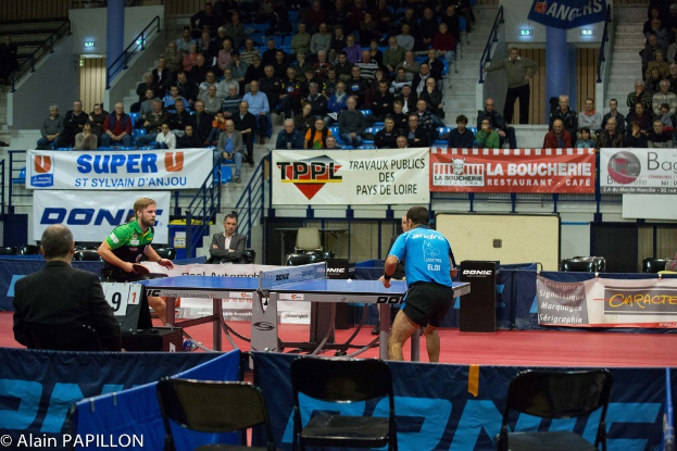 Indoor-Tischtennis-Match mit spielenden Spielern, Zuschauern im Sitzen und Stehen, leeren Stühlen und Werbeplakaten im Hintergrund.