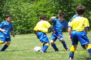 Kinder spielen Fußball auf einem Rasenplatz mit Bäumen im Hintergrund und einem Datumsstempel in der rechten unteren Ecke.