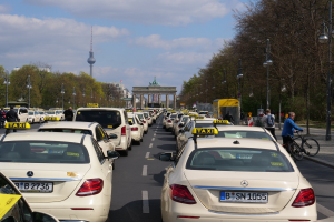 Eine lange Reihe von Taxis steht an einer belebten Straße in Berlin, Deutschland, mit Fahrradfahrern und Fußgängern auf dem Gehweg, flankiert von Bäumen und Laternen, und Gebäuden, einem Bogen und einem Turm im Hintergrund unter einem bewölkten Himmel.