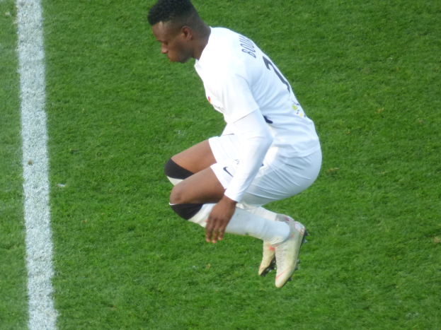 A professional soccer player in a white uniform kneels on a soccer field, wearing football boots.