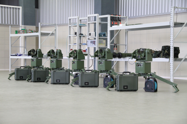 Eine Gruppe von Militärrobotern auf einem Boden mit Koffern und Gegenständen auf Gestellen, positioniert in der Nähe einer Wand und eines Geländers als Teil eines Verteidigungssystems.