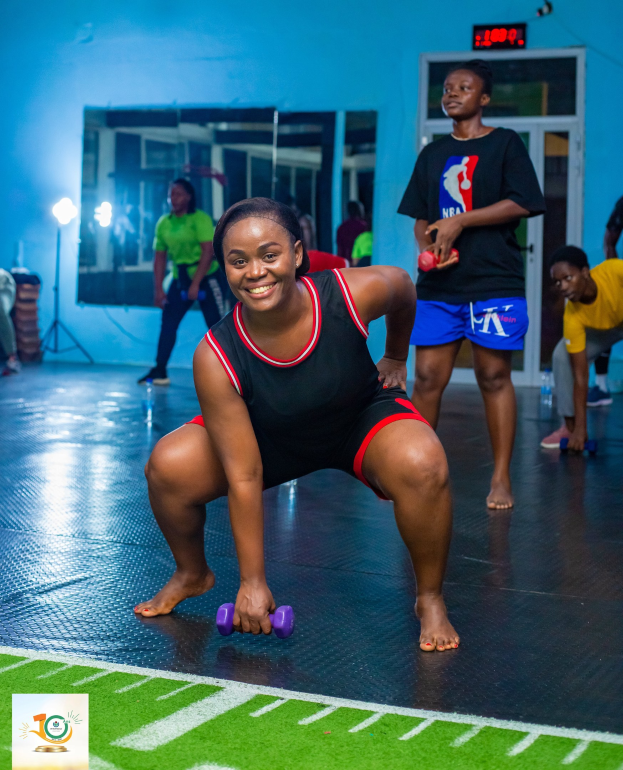 Eine Frau in Kniebeugeposition mit Hanteln in einem Fitnessstudio mit Equipment und Logos im Hintergrund.