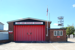 Feuerwehr- und Rettungsdienstgebäude mit roter Tür, Fenstern, einem Namensschild, einem Fahnenmast mit Flagge, einem Metallturm, einem Zaun, einer Gruppe von Bäumen und einem bewölkten Himmel.