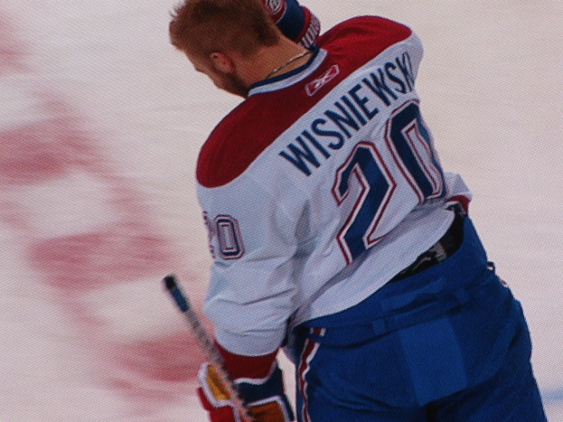 Ein Hockey-Spieler in einem "Wisniewski"-Trikot steht auf dem Eis, hält einen Hockey-Schläger und hat einen konzentrierten Gesichtsausdruck.