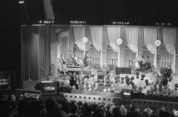 Schwarzes und weißes Foto einer Gruppe von Menschen auf einer Bühne mit Ballons und Fernsehern im Hintergrund.