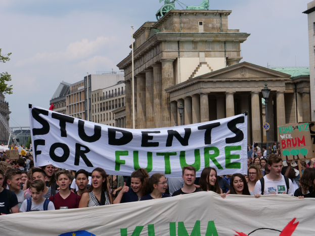 Gruppe von Schülern marschiert in Berlin mit einer leuchtend bunten "Students for Future"-Schultertasche vor einem Hintergrund aus Gebäuden, Bäumen und Himmel.