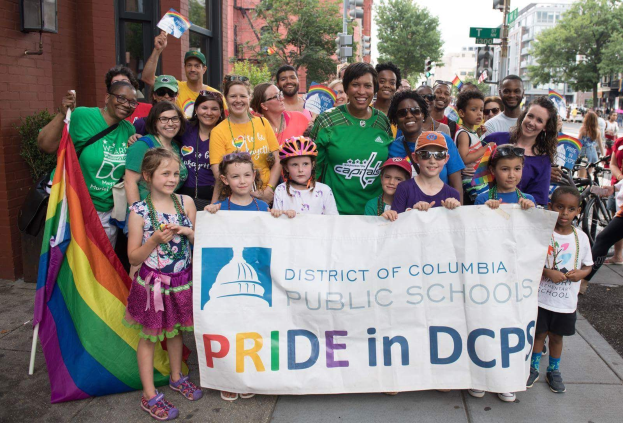 Gruppe von Menschen auf dem Boden, einige halten ein Banner mit der Aufschrift "Bezirk Columbia Public Schools Pride in DCPS", mit Kindern, die eine Fahne halten, einer Person auf einem Fahrrad, Bäumen, Gebäuden, Straßenschildern, Ampeln und einem bewölkten Himmel.