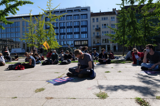 Eine Gruppe von Menschen sitzt vor einem Gebäude mit Bäumen und einem klaren blauen Himmel auf dem Boden, einige tragen Masken, mit verstreuten Taschen und Gegenständen, in einer Protestszene in Berlin.