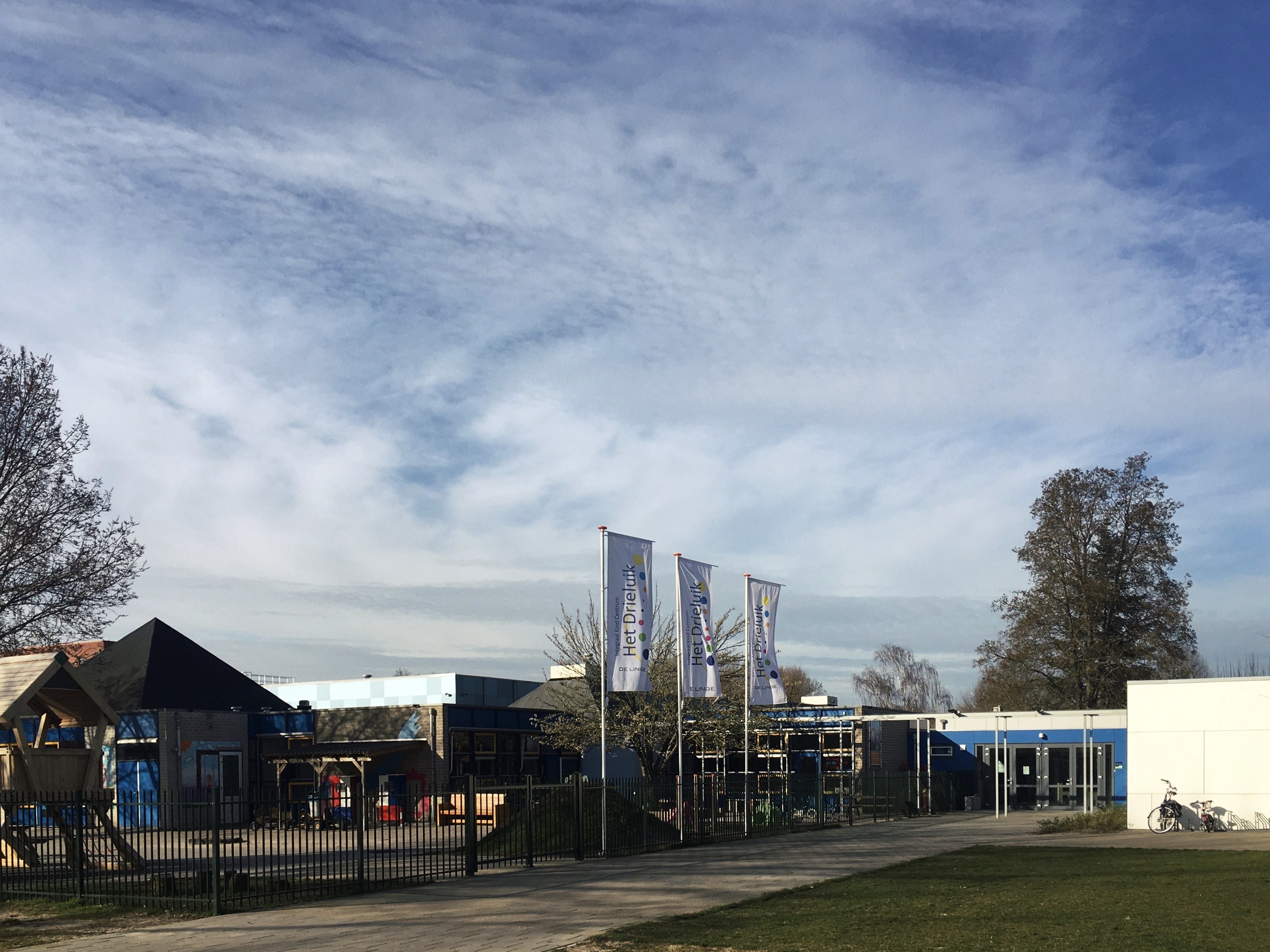 Großes Gebäude mit einem grasigen Bereich davor, umgeben von einem Metallzaun, mit einem Weg, Bäumen, Pfosten, Bannern, einem geparkten Fahrrad, nahen Häusern und einem bewölkten Himmel, als neuer Spielbereich für Kinder an der Schule identifiziert.